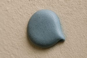 Close-up of Blu Tack blob on a textured painted wall, revealing tiny air bubbles, paint cracks, and discoloration. Captured with Canon EOS 5D Mark IV, macro lens. Soft lighting highlights natural imperfections in Blu Tack's adhesion and varied wall textures.