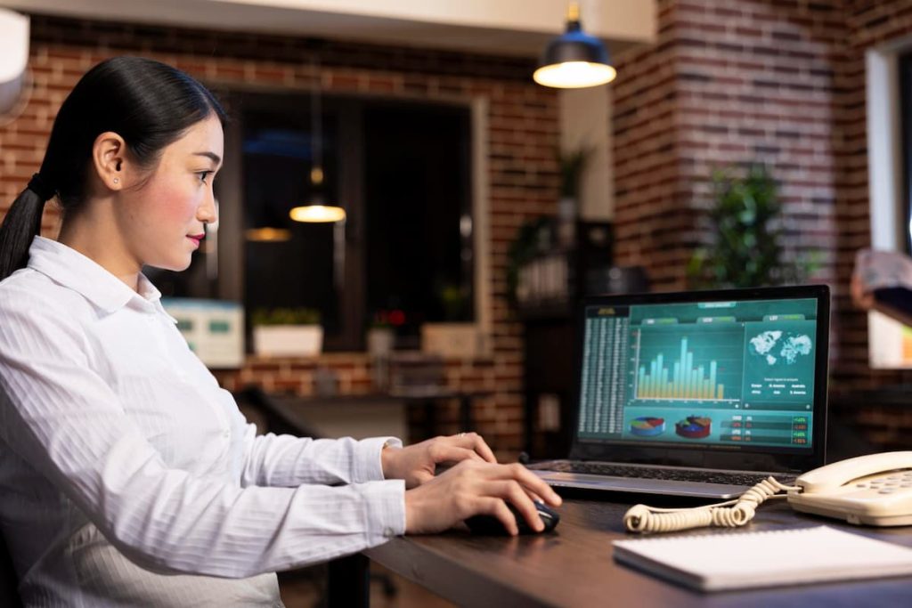 A woman in business attire sits at a desk working on a laptop displaying charts and graphs, focused on real-time scheduling to avoid delays in a modern office setting with brick walls and plants.