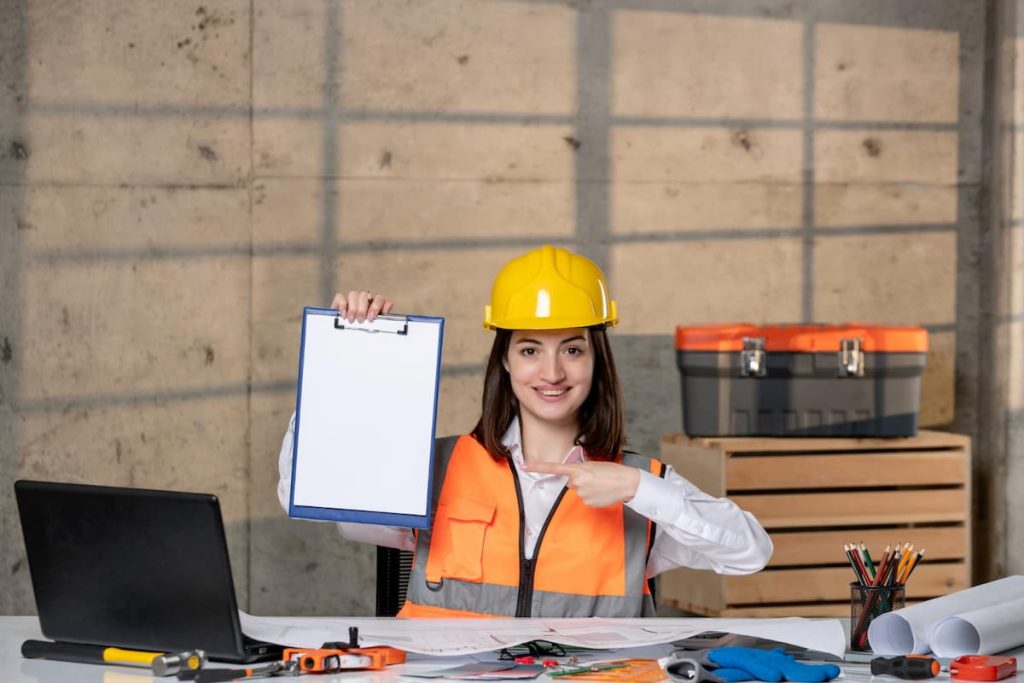 A person in a hard hat and safety vest sits at a desk with tools and plans, holding up and pointing to a blank clipboard—ready to outline the next steps using advanced digital construction scheduling tools.