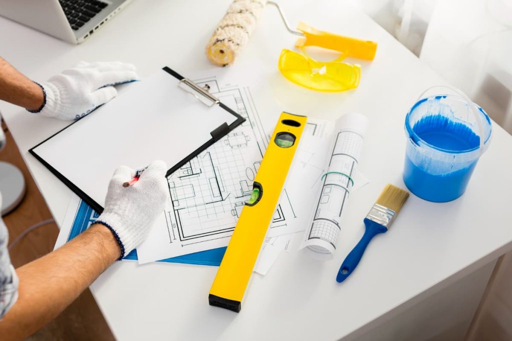 Person wearing gloves writes on a clipboard over architectural plans, highlighting key steps in the construction process, with a spirit level, paintbrush, blue paint, safety spectacles, and a paint roller on a white table.