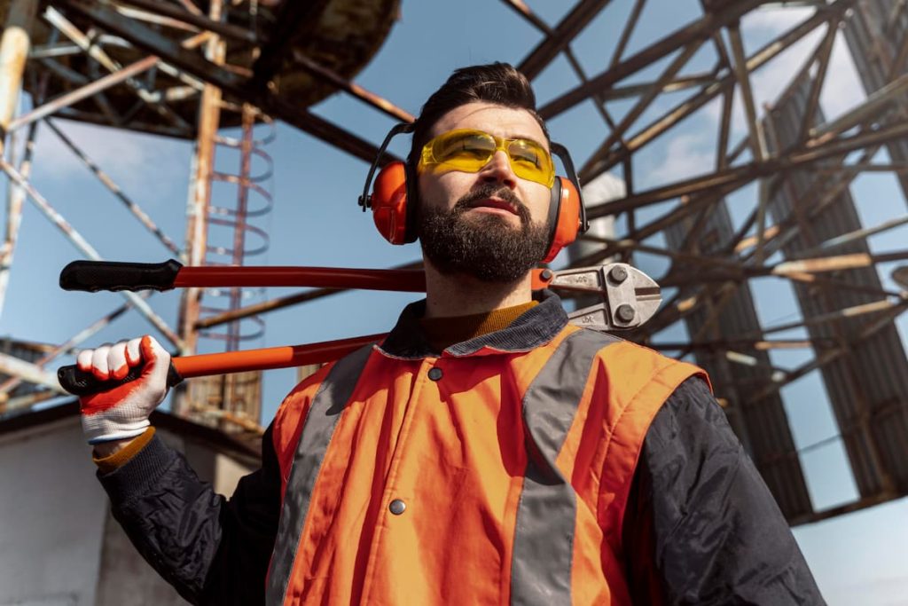 A construction worker wearing safety gear and holding bolt croppers over his shoulder stands outdoors at an industrial site with metal structures in the background, reflecting the dedication of overseas workers employed by leading construction companies as they prepare for projects launching in 2026.