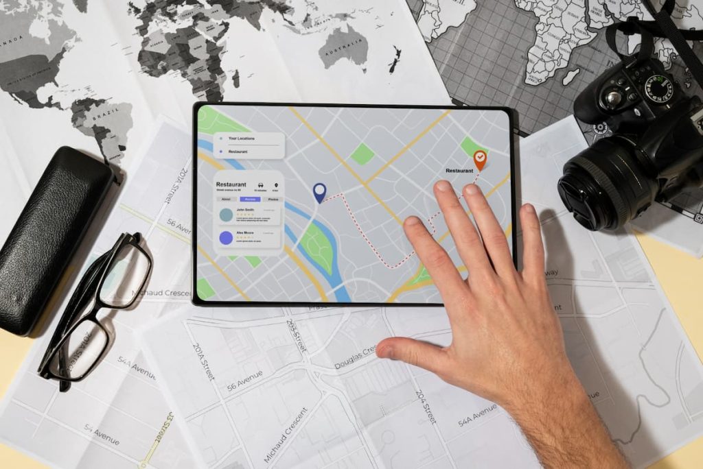 A hand touches a tablet displaying a digital map with restaurant locations, optimising local SEO for better visibility; maps, spectacles, a camera, and a case are arranged around it to help generate enquiries for local trades.