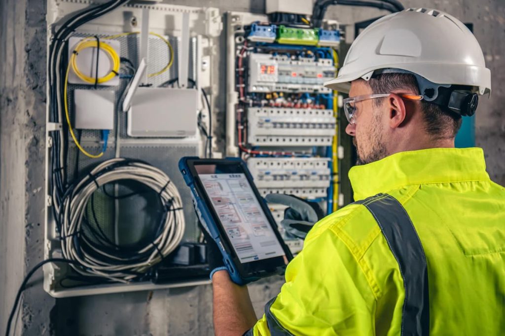 An electrician wearing a hard hat and high-visibility jacket inspects an open electrical panel whilst using a tablet for data entry or diagnostics, ensuring thorough electrical safety checks for property owners in accordance with EICR London regulations.