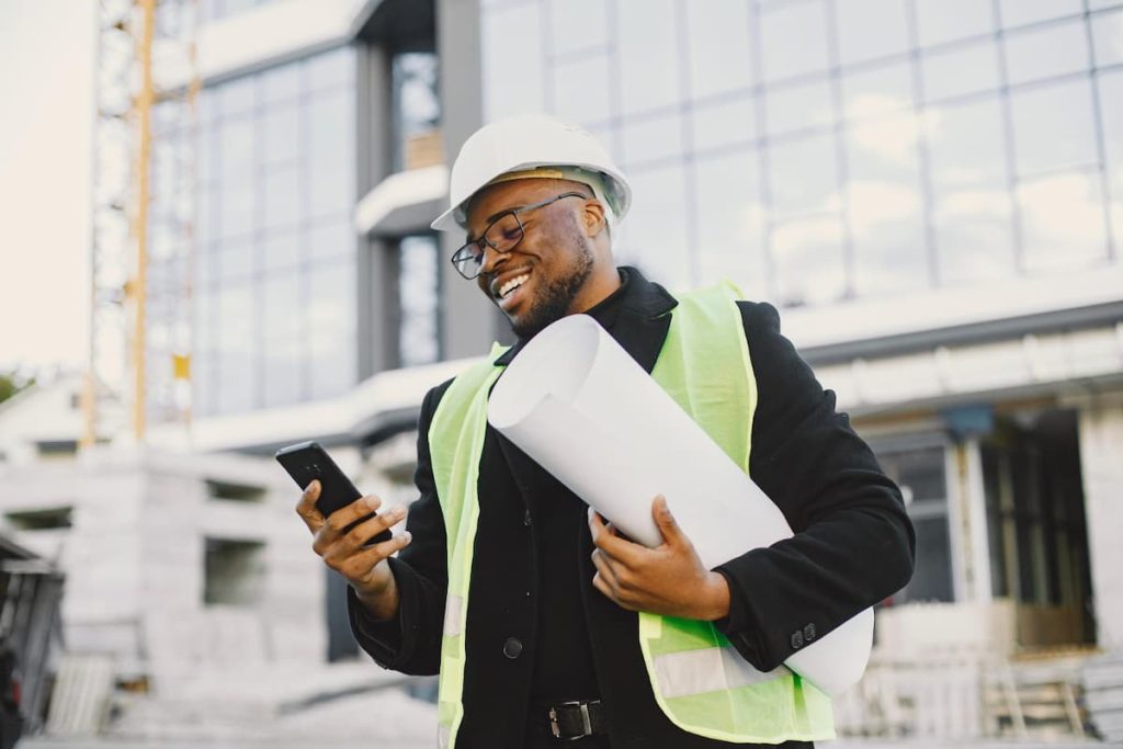 A man in a safety vest and hard hat holds plans and smiles at his mobile on a building site with a modern glass building in the background, possibly reviewing contractor estimates or managing construction payments.