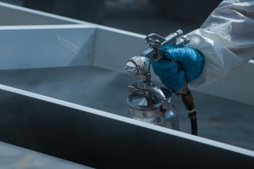 A person wearing blue gloves and protective clothing uses a spray gun to apply metal coatings to a surface, providing essential corrosion protection in construction environments.
