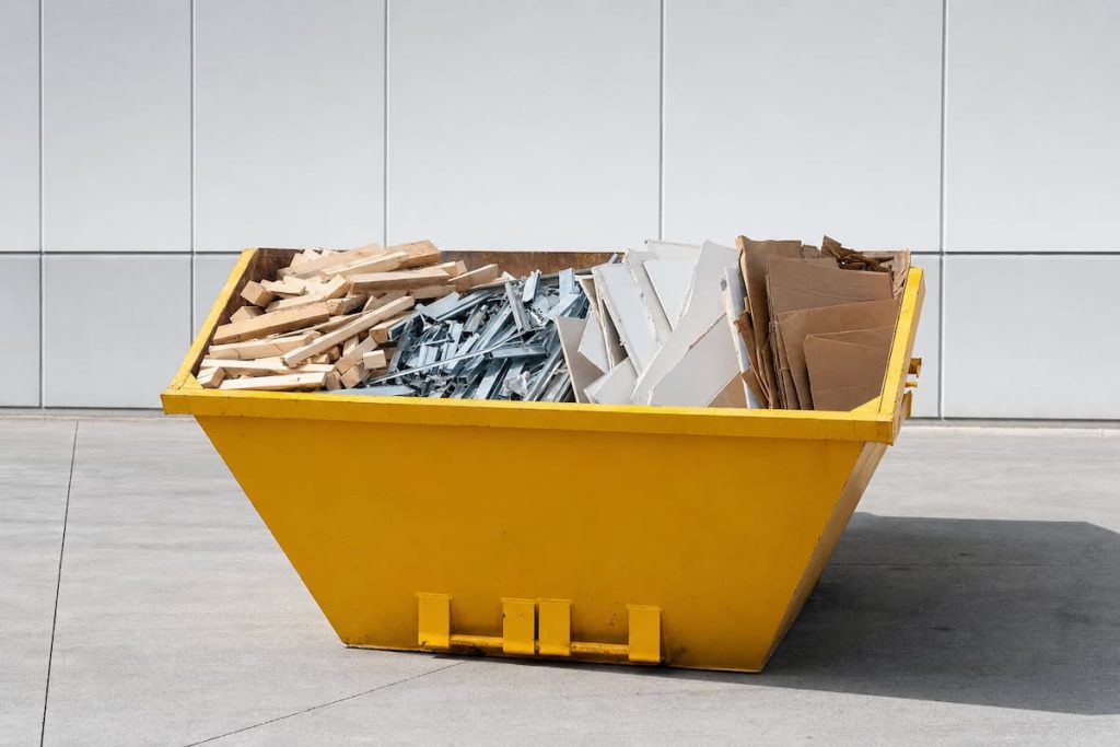 A yellow skip on concrete holds wood, metal, and cardboard debris in front of a white tiled wall, reflecting the sustainable waste practices increasingly adopted by the UK construction industry.