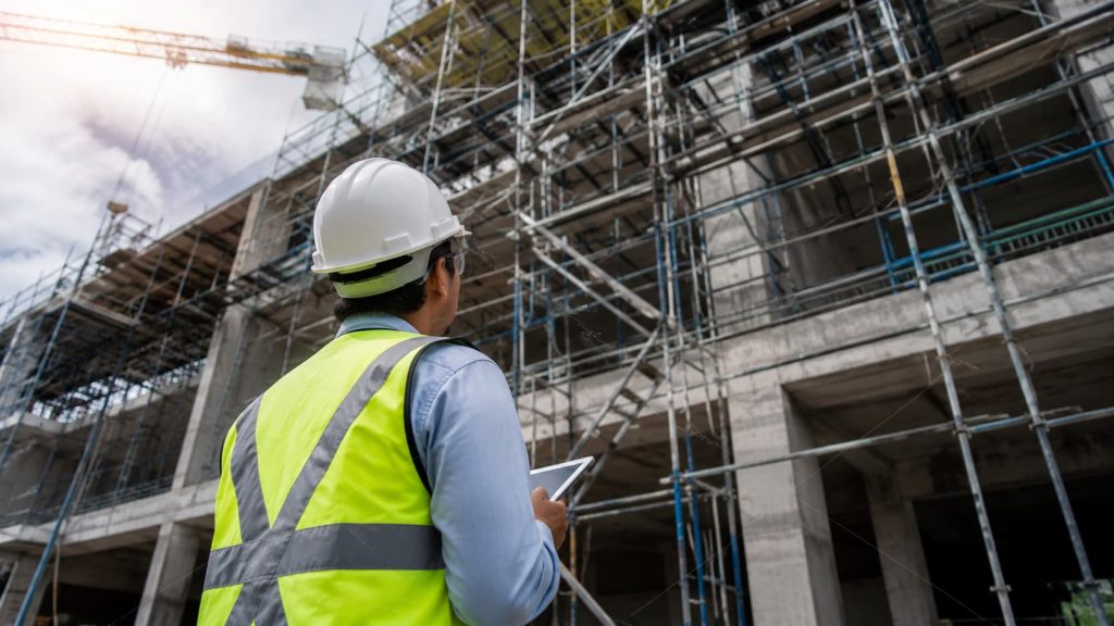 A construction worker in a hi-vis vest and hard hat stands in front of scaffolding, holding a tablet and looking up at a building under construction, overseeing the latest delivery from Westline acquisition for Wade Building Services’ building supply needs.