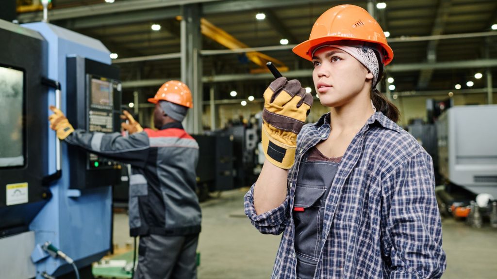 Two factory workers wearing safety gear operate machinery; one speaks into a walkie-talkie whilst the other uses a control panel in an industrial setting, demonstrating the importance of healthy work environments often emphasised in the construction industry.
