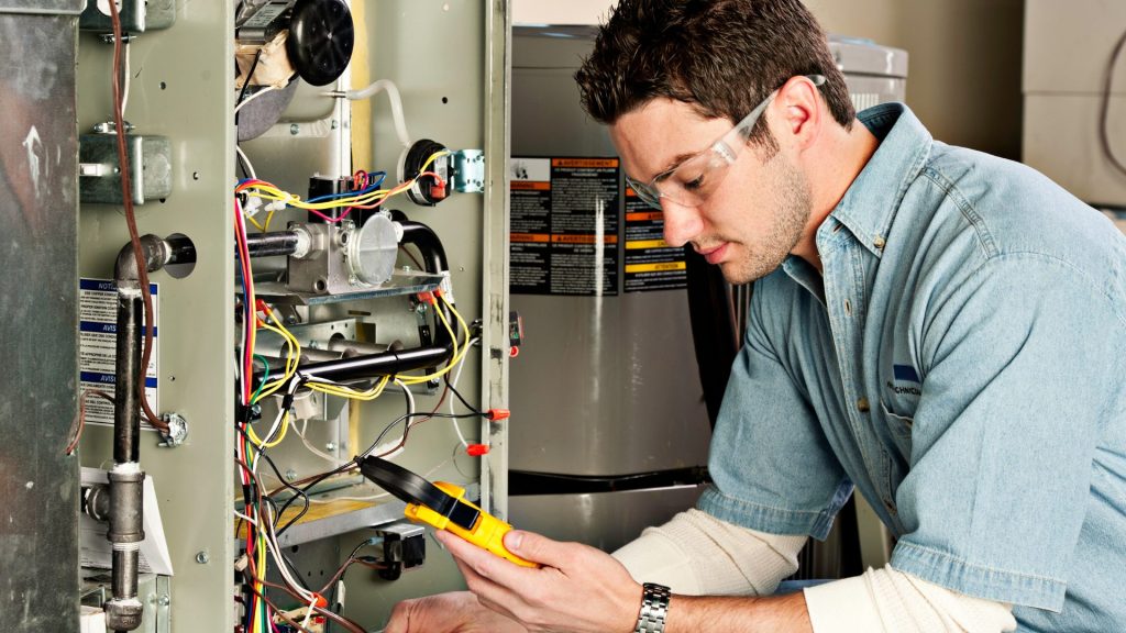 A technician wearing safety spectacles uses a multimeter to check electrical components inside an open furnace unit, carefully inspecting for common furnace signs that may indicate the need for furnace repair or help prevent a potential furnace breakdown.