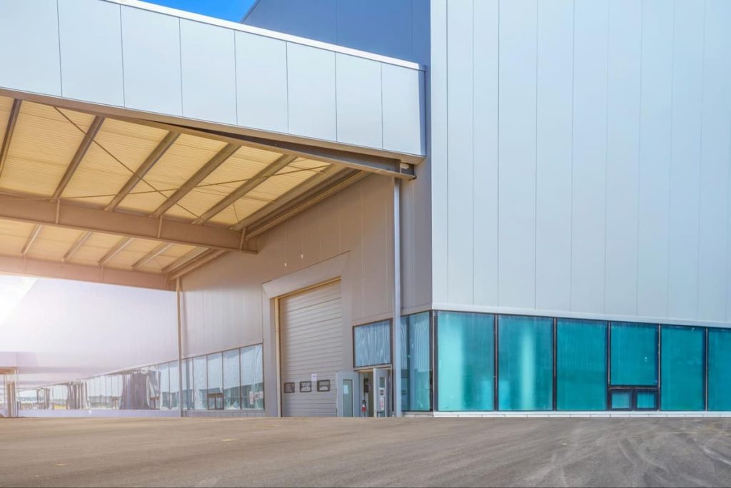Large industrial warehouse with a partially open roller shutter door, tall metal walls, blue-tinted windows, and an empty tarmac area in front—designed with expertise in UK Cold Storage Construction for optimal efficiency and temperature control.