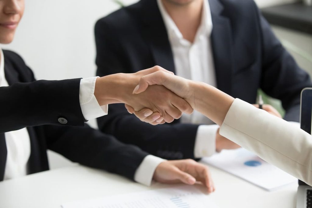 Two people in business attire shake hands across a desk whilst a third person sits nearby with documents, suggesting a business meeting or agreement focused on boosting field team efficiency in the UK.