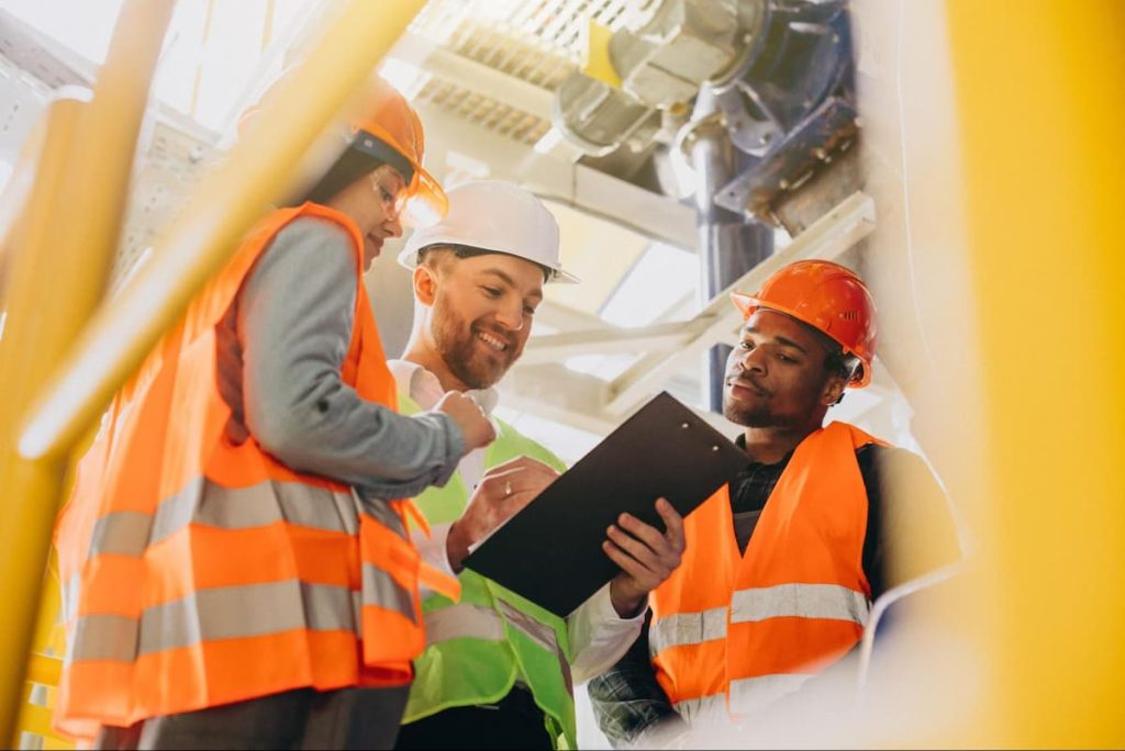 Three construction workers wearing safety gear and hard hats review a clipboard together at a worksite, ensuring all pre-refurbishment surveys are completed before contractors begin the refurbishment.