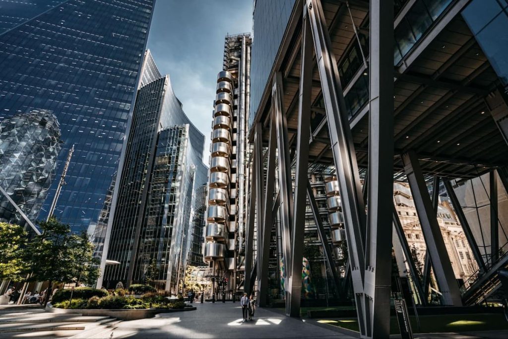 Modern cityscape with tall, reflective glass buildings, a metallic cylindrical structure, and people walking in a sunny urban environment—captured through high-quality 3D exterior design rendering to showcase the vibrancy and architectural innovation of new development tenders.