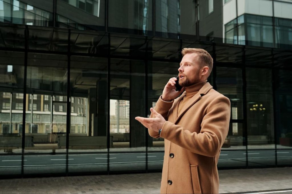 Man in a tan coat stands outside a glass office building, talking on a mobile phone and gesturing with his left hand—demonstrating how UK field teams boost productivity through effective communication.