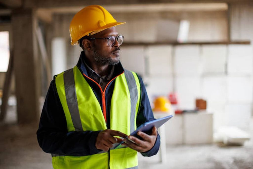 Man wearing a yellow hard hat and reflective vest holds a tablet at a building site, with building materials in the background—demonstrating how DDA inspectors help businesses maintain compliance and prevent legal action.