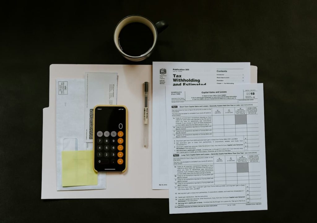 A folder with tax documents, a pen, a smartphone calculator, envelopes, sticky notes, and a cup of coffee are arranged on a dark surface—an organised workspace that reflects the attention to detail needed for tax compliance in construction companies.