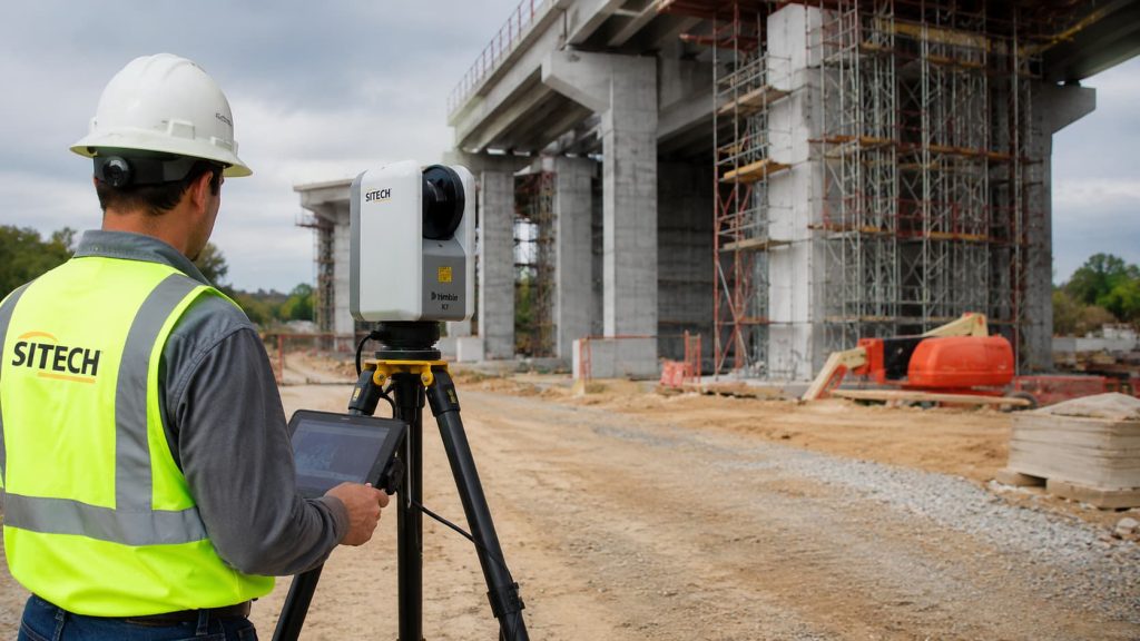 A construction worker in a hard hat and safety vest uses a surveying instrument near a partially built bridge with scaffolding and equipment visible, demonstrating the importance of precise mobile mapping on UK construction sites.