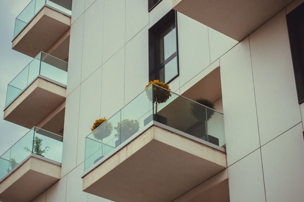 Modern apartment block with glass balustrades on each balcony; each balcony has potted plants, including yellow-flowered shrubs. The facade is white with dark window frames, creating light-filled spaces throughout the building.
