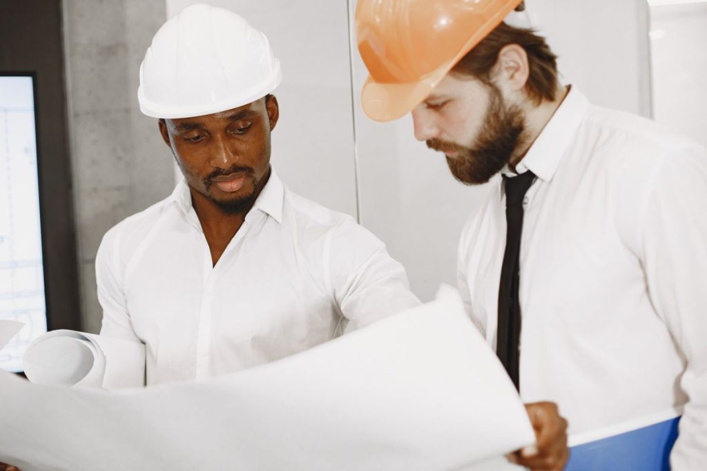 Two men in hard hats and white shirts review a large blueprint together at an international construction site, carefully checking for any translation errors that might have occurred in the technical manuals.