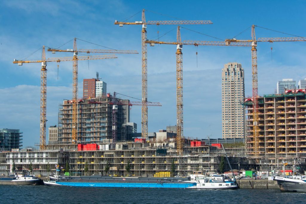 Several construction cranes operate above buildings under construction near a waterfront in a city, where new water connections and contestable split works are underway on site, with high-rise buildings visible in the background under a blue sky.