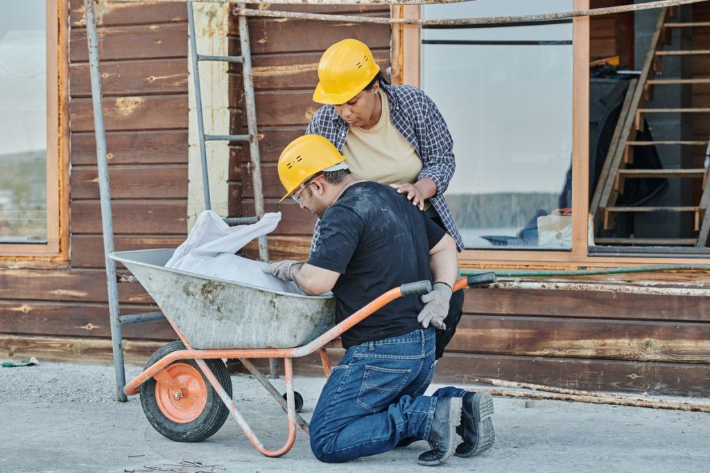 Two construction workers wearing yellow helmets examine materials in a wheelbarrow outside a building under construction, staying vigilant to avoid potential workplace injury.