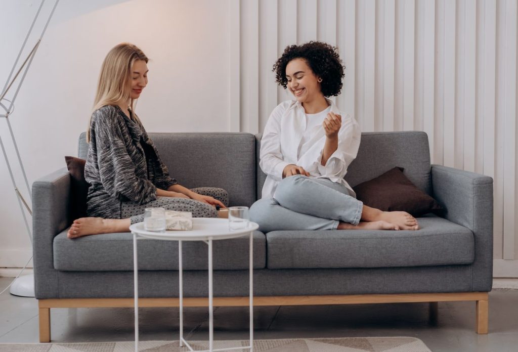 Two women sit barefoot on a grey FCI London sofa in a modern living room, talking and smiling. A small round table in front of them holds glasses of water and a bowl, creating an inviting space that reflects the stylish touch of Interior Designers London.