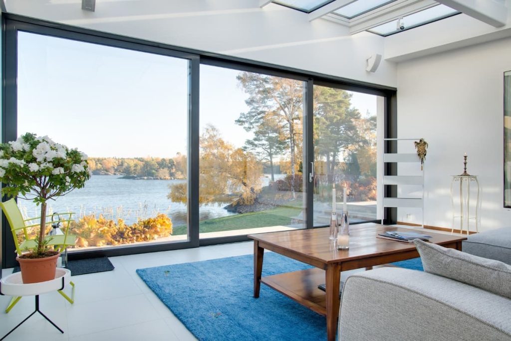 Modern living room with large glass doors boasting excellent sliding door performance, overlooking a lake and autumn trees, featuring a blue rug, wooden coffee table, grey sofa, and indoor plant.