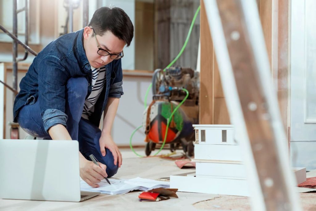 A person kneels on the floor, reviewing papers and writing, with a laptop and architectural model nearby in a construction setting—carefully considering plumbing inspection notes whilst renovating an older home.