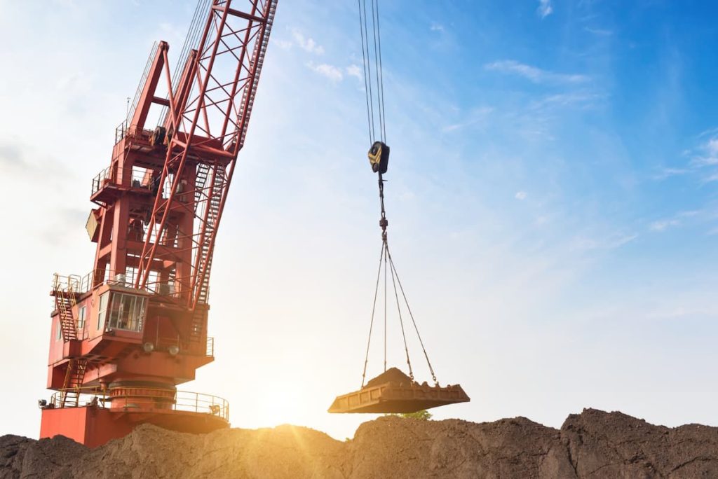 A large red crane utilises vertical transport to lift a heavy load of earth or material against a bright blue sky with sunlight in the background.