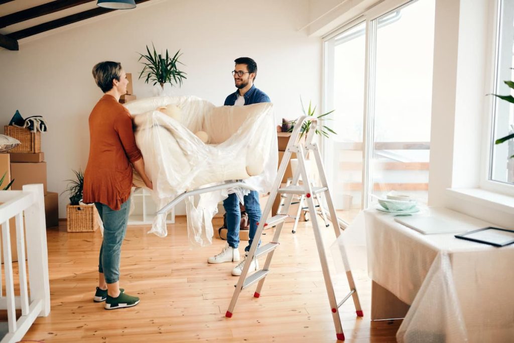 Two people move a wrapped armchair in a bright room with wooden floors, plants, boxes, and a stepladder nearby—capturing the busy atmosphere often seen during post-renovation cleaning.