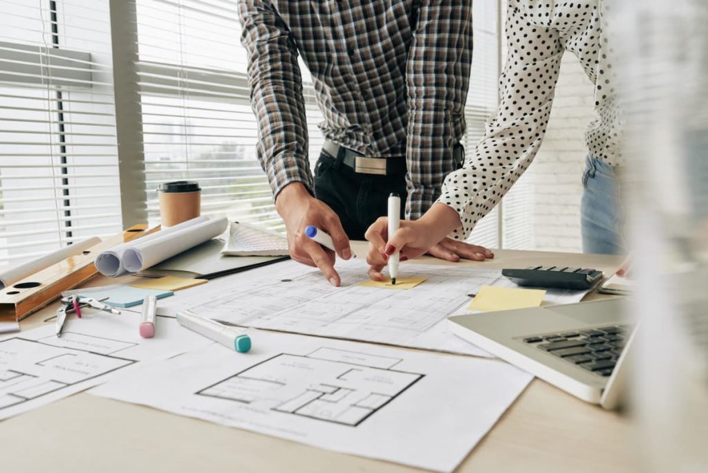 Two people review and mark up architectural blueprints on a desk with a laptop, calculator, coffee cup, and rolled-up papers nearby, ensuring site data accuracy throughout their planning process.