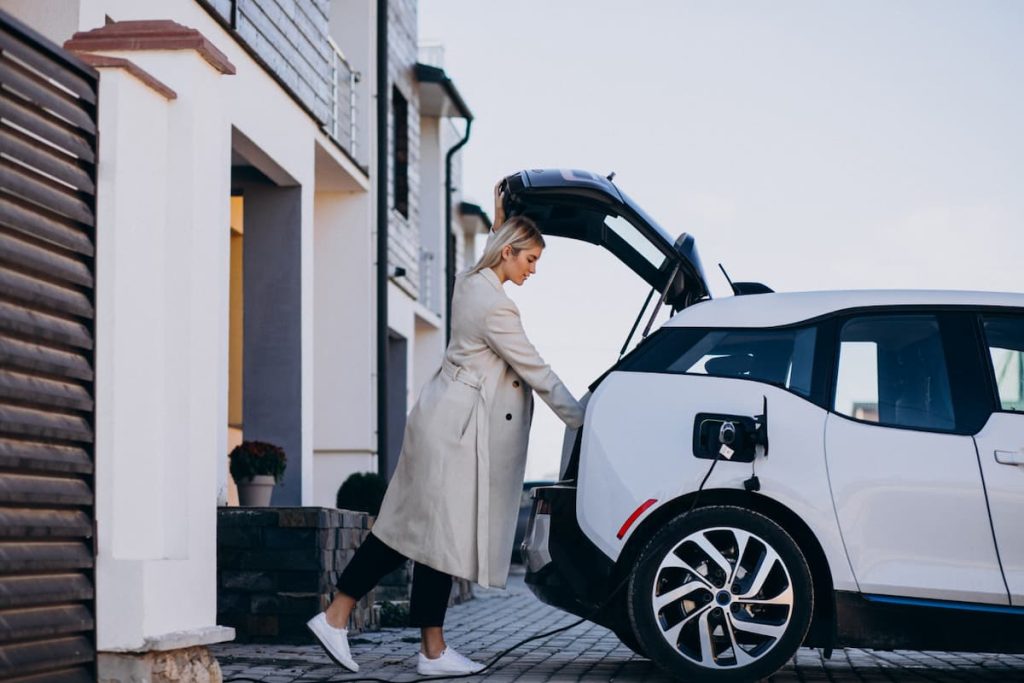 A person in a light coat stands next to a white electric car with an open boot and charging cable, outside a modern house, highlighting the convenience of home EV charging for drivers.