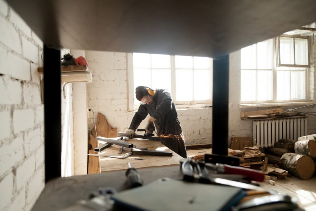A person wearing safety gear uses a power tool to cut metal in a workshop with exposed brick walls, wooden logs, and scattered tools—capturing the hands-on process often seen during the full renovation of an older property or its HMO conversion.