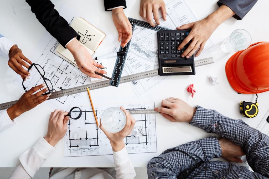 Overhead view of people collaborating on architectural blueprints, using a calculator, ruler, pencil, and water bottle, with a red hard hat and measuring tape nearby—illustrating how effective construction project management can help bridge the site-to-office gap.