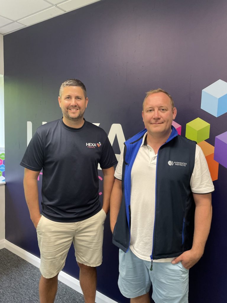 Two men standing indoors in front of a dark wall with colourful cube designs, both smiling at the camera, celebrate the success of Hexa Finance’s latest construction campaign.