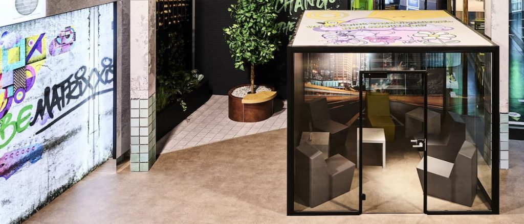 Glass meeting pod with five chairs inside, situated in a modern indoor space featuring graffiti wall art and potted plants nearby—a stylish setting that perfectly complements sustainable exhibition stands and contemporary exhibition stand construction.