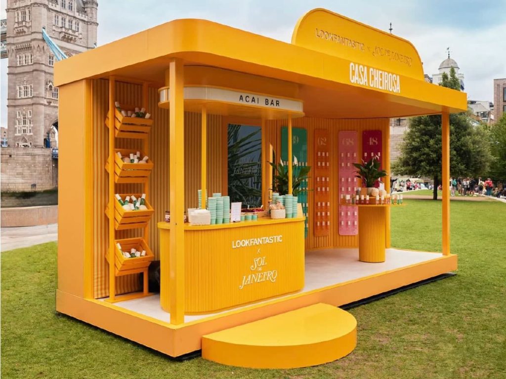 A bright yellow pop-up stall labelled “Casa Cheirosa” displays beauty products on shelves and counters, showcasing innovative exhibition stand construction against a backdrop of lawn and historic buildings.