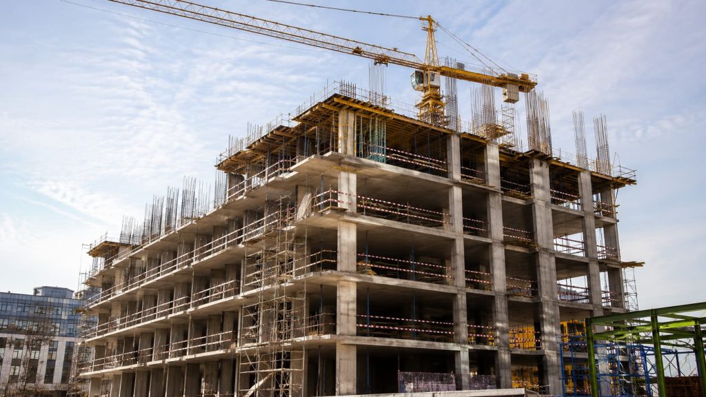 A multi-storey concrete building under construction with scaffolding, a yellow tower crane, and exposed steel reinforcements under a partly cloudy sky, where careful management of construction timetables is essential to meet project goals.