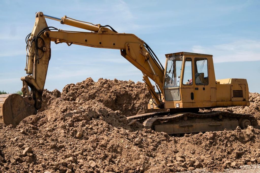 Yellow Yanmar powered excavator positioned on a large mound of earth under a blue sky, with its bucket—featuring genuine parts—partially submerged in the soil.