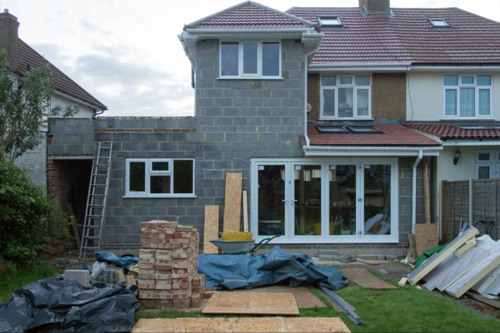 - Machinery Asia A house under construction with grey brick walls, building materials, and debris scattered in the garden; ladders and tools are visible.