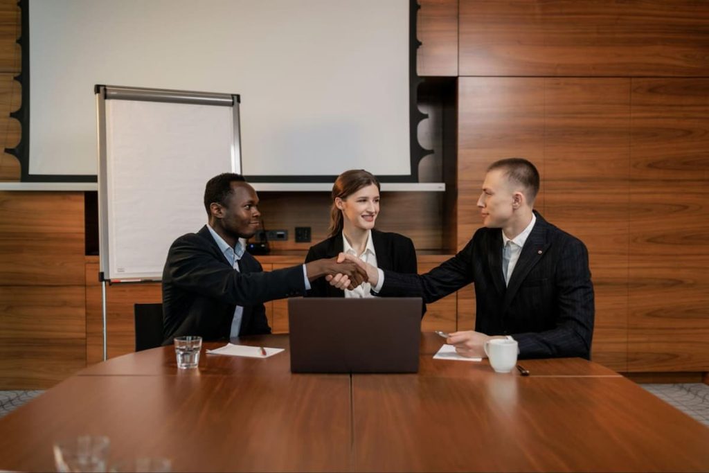 Three people in business attire sit at a conference table; two men, likely property owners or representatives of a professional property management firm, shake hands while a woman looks on. A laptop, papers, and cups are on the table, suggesting a productive meeting about property management, with a flip chart in the background.