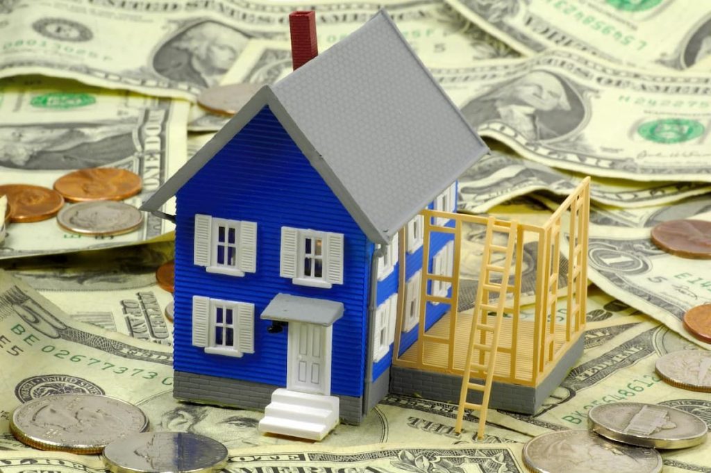 A model house with a partially built extension sits on top of assorted US dollar notes and coins, symbolising home construction and financial investment—much like the careful consideration needed during a home inspection to spot potential problems before viewing a home.