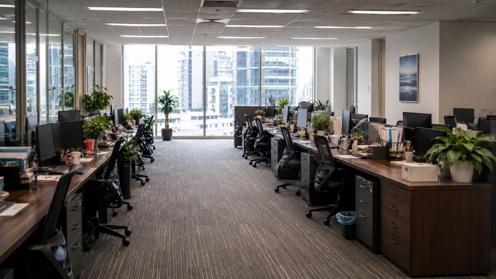 Modern office space with rows of desks, computers, chairs, and plants. Large windows at the end show tall buildings outside, offering a prime workplace location to compare with other offices. The office appears empty and well-organised—a setting ideal for founders seeking an inspiring environment.