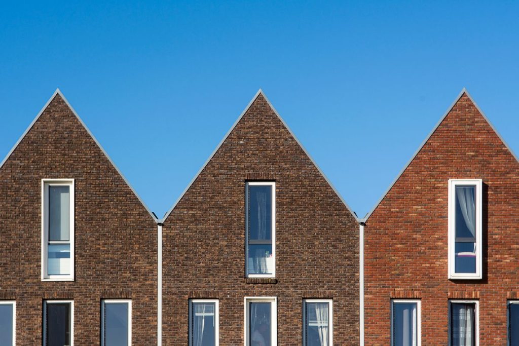 Three modern houses with steep triangular roofs, large vertical windows, and brick façades stand side by side against a clear blue sky—a testament to the efficiency of UK contractors renowned for delivering fast-track builds.