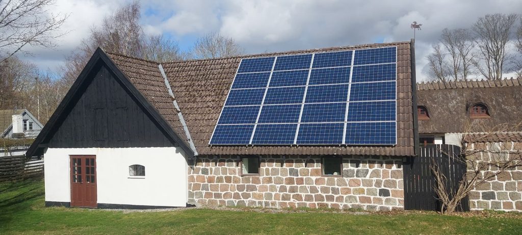 A stone and white house with a sloping roof has large solar panels installed on one side, surrounded by grass and trees under a partly cloudy sky—a scene that’s becoming increasingly common for UK homeowners looking to embrace clean energy by 2026, despite lingering solar panel myths.