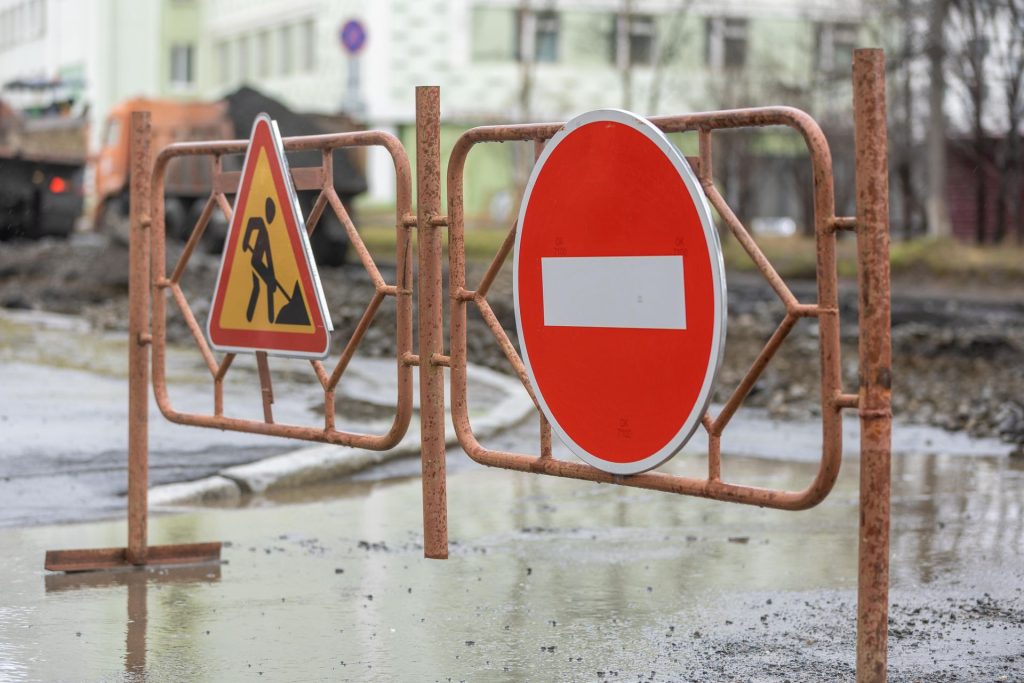 Roadworks and no entry construction signs are displayed on metal barriers blocking a wet road, with construction activity and buildings visible in the background. The distinct construction sign colours help convey the meaning of construction signs, ensuring drivers and pedestrians recognise hazards and follow safety instructions.