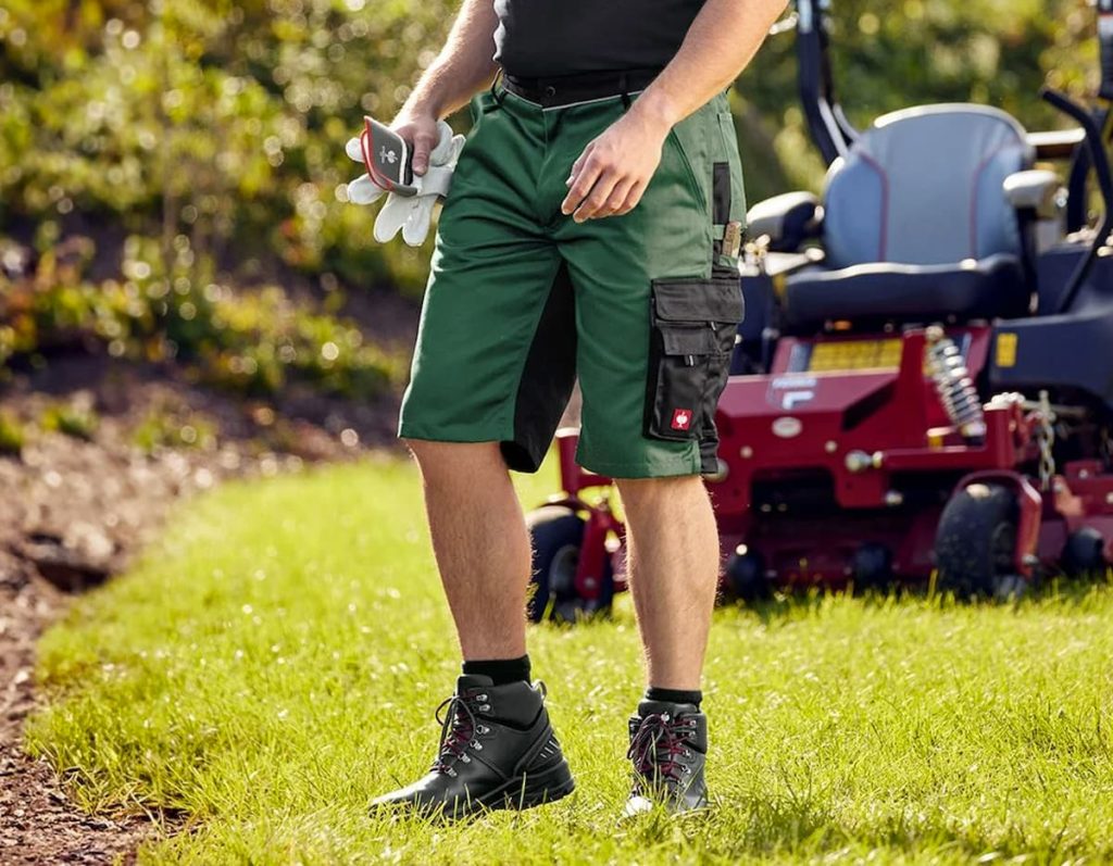Person in durable work shorts and black boots stands on grass, holding gloves, with a red lawnmower in the background—ready for garden work with summer comfort and safety in mind.