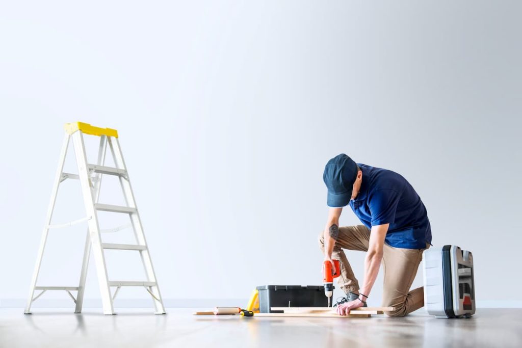 A person in a blue shirt kneels on the floor using a power drill on wood. Toolbox, tools, and a ladder are nearby against a plain white wall, with visible dust after renovation still lingering throughout the workspace.