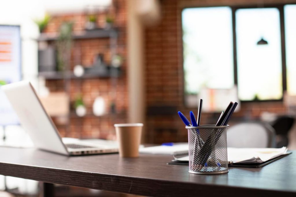 A desk with an open laptop, a paper cup, pens in a metal holder, and documents creates an organised workspace that enhances workplace performance. The background reveals a window and shelves with decorations, highlighting the importance of thoughtful office furniture in a productive environment.