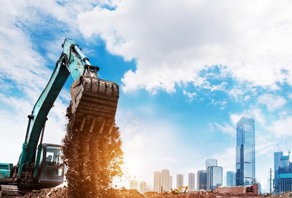 An excavator dumps soil at a construction site with a city skyline and tall buildings in the background under a partly cloudy sky, as part of Net Zero initiatives requiring careful verification of sustainable building practices.